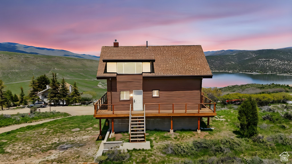 Back of house at dusk with a deck with water view, a chimney, roof with shingles, and stairway