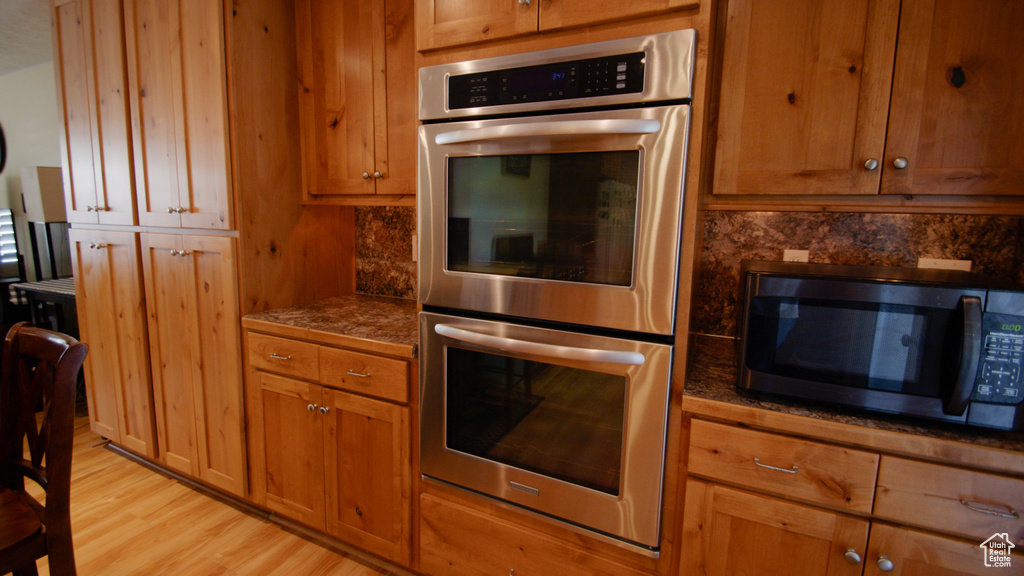 Kitchen with stainless steel double oven, light wood-style flooring, brown cabinets, and dark countertops