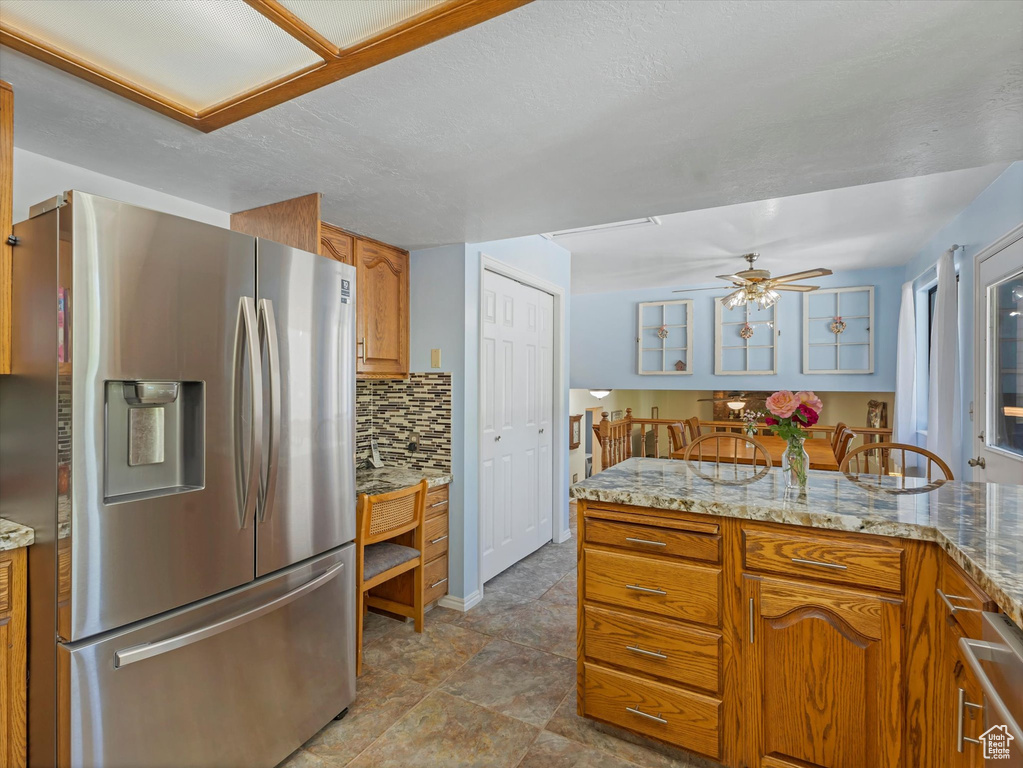 Kitchen with stainless steel refrigerator with ice dispenser, decorative backsplash, ceiling fan, brown cabinets, and light stone counters