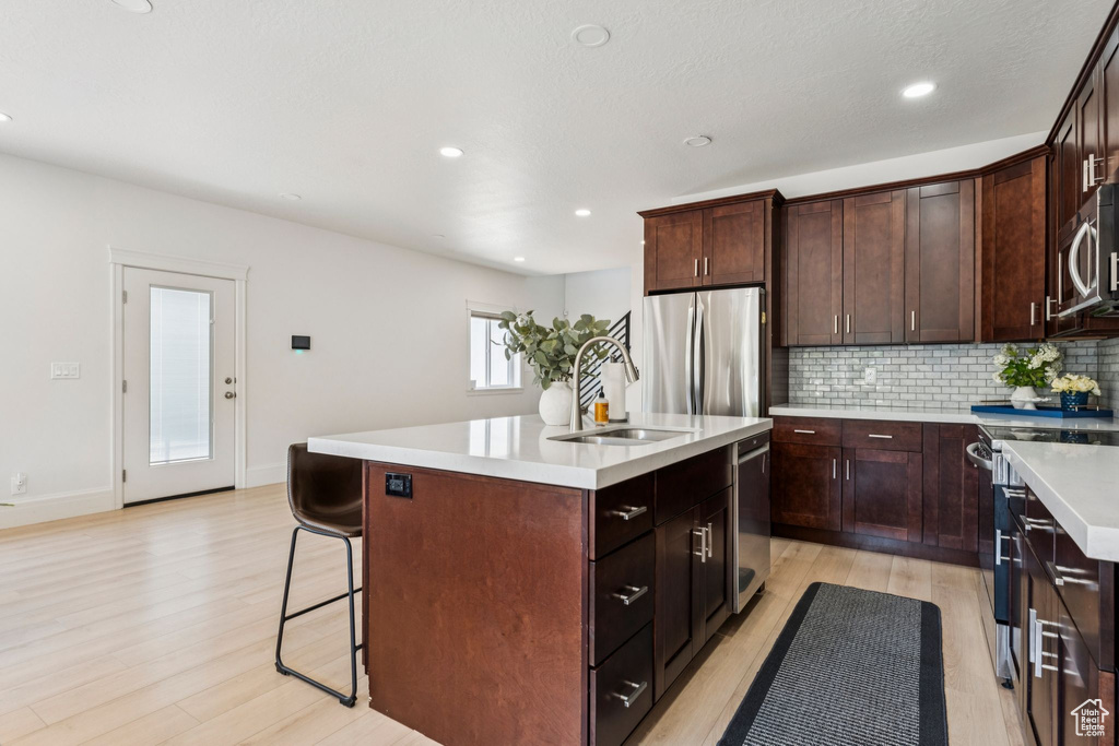 Kitchen featuring appliances with stainless steel finishes, light wood finished floors, a center island with sink, decorative backsplash, and light countertops