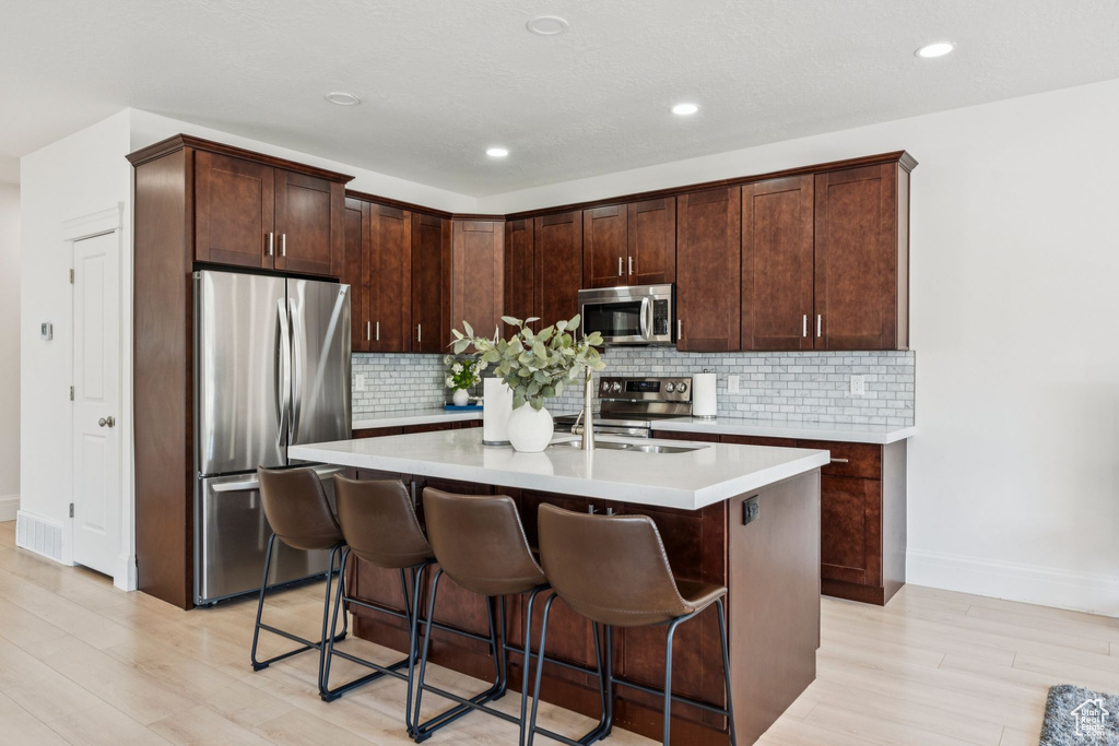 Kitchen featuring stainless steel appliances, light wood finished floors, light countertops, a kitchen bar, and a kitchen island with sink