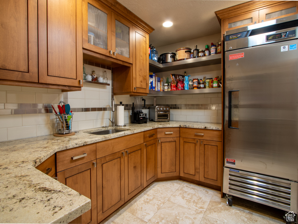 Kitchen featuring stainless steel refrigerator, brown cabinets, decorative backsplash, light stone counters, and glass insert cabinets