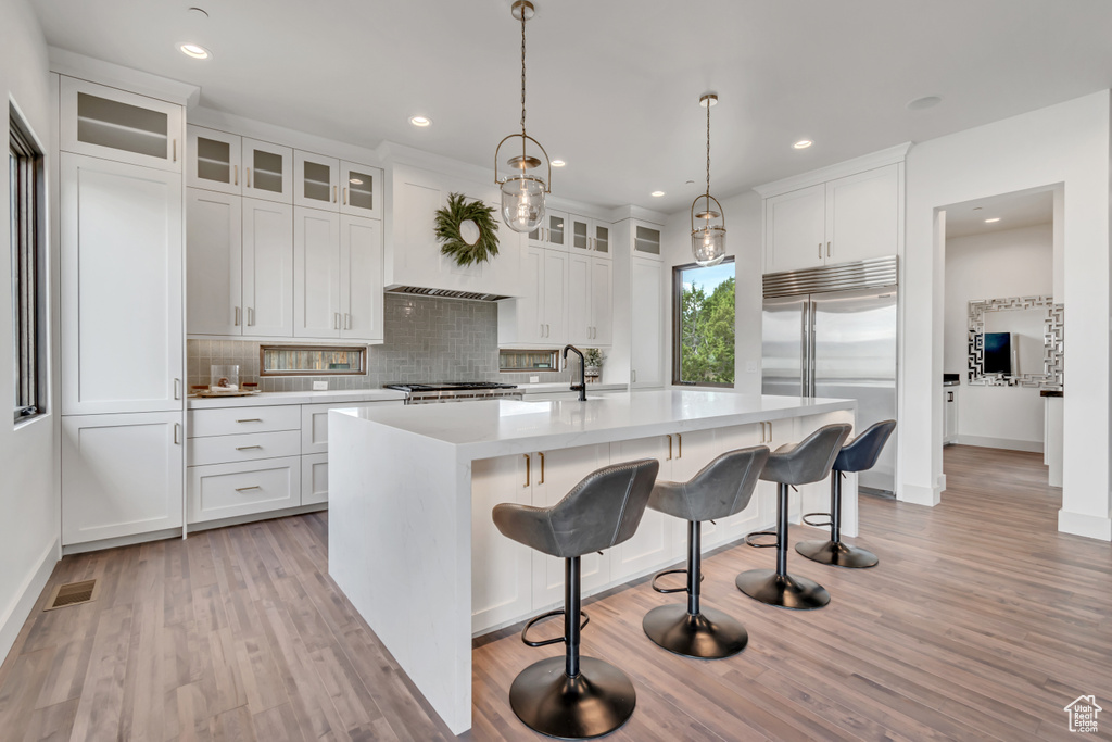 Kitchen featuring stainless steel built in refrigerator, light wood-type flooring, decorative backsplash, light countertops, and an island with sink