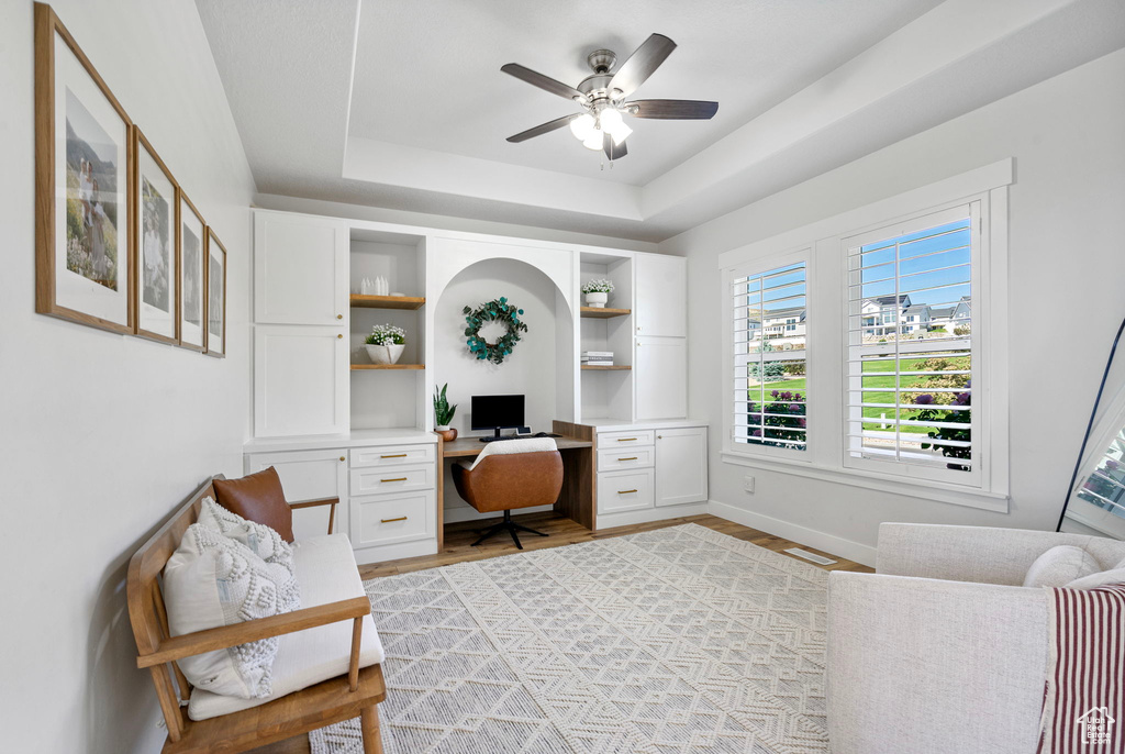 Office space featuring a ceiling fan, baseboards, a raised ceiling, built in study area, and light wood-style flooring