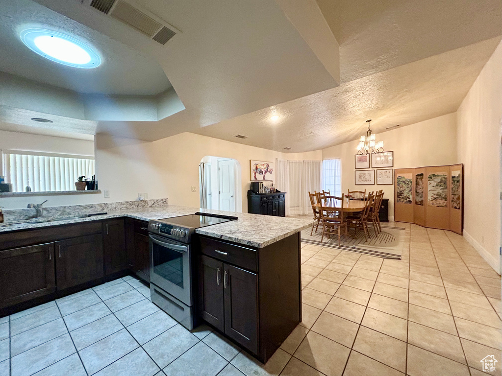 Kitchen with stainless steel range with electric stovetop, a peninsula, light tile patterned flooring, light stone counters, and a chandelier