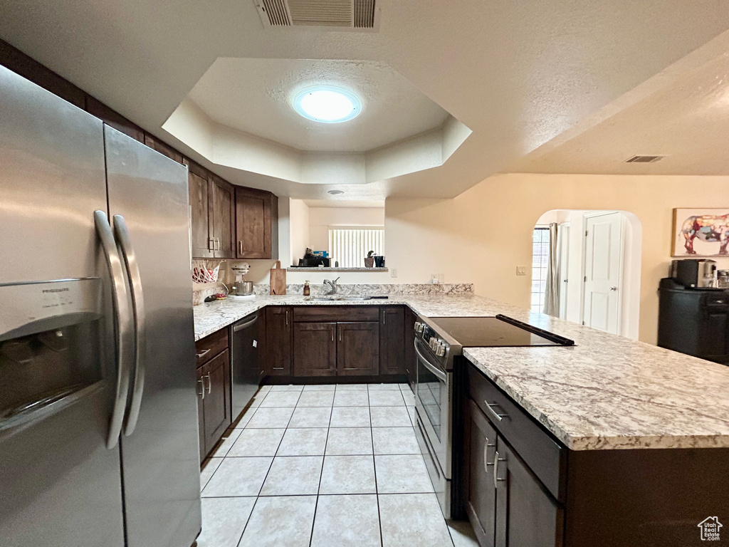 Kitchen featuring appliances with stainless steel finishes, a peninsula, dark brown cabinets, light tile patterned floors, and a raised ceiling