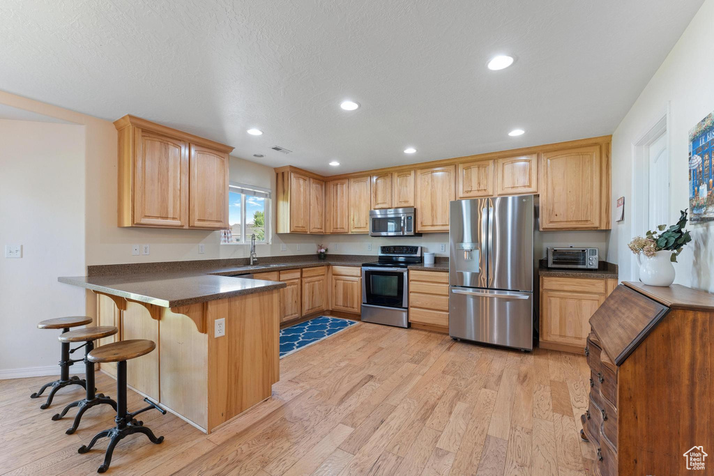 Kitchen featuring appliances with stainless steel finishes, a peninsula, light wood finished floors, dark countertops, and recessed lighting