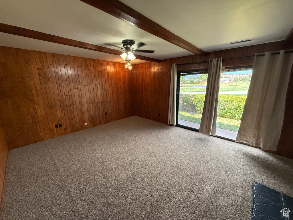 Carpeted spare room featuring wood walls, beamed ceiling, and a ceiling fan