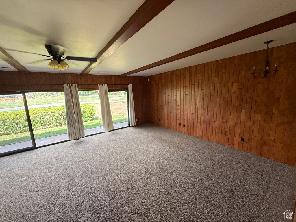 Carpeted empty room with wooden walls, beamed ceiling, ceiling fan, and a chandelier