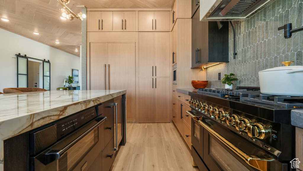 Kitchen with range with two ovens, wall chimney exhaust hood, light wood-type flooring, light stone counters, and backsplash