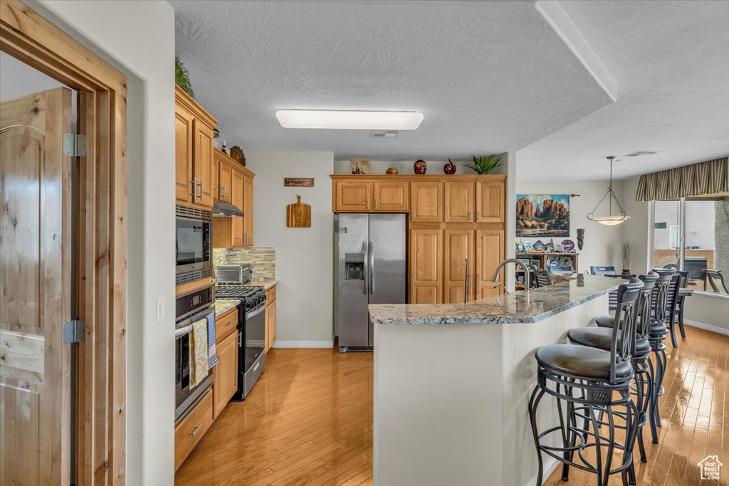 Kitchen featuring appliances with stainless steel finishes, a kitchen breakfast bar, light wood-type flooring, a textured ceiling, and light stone countertops