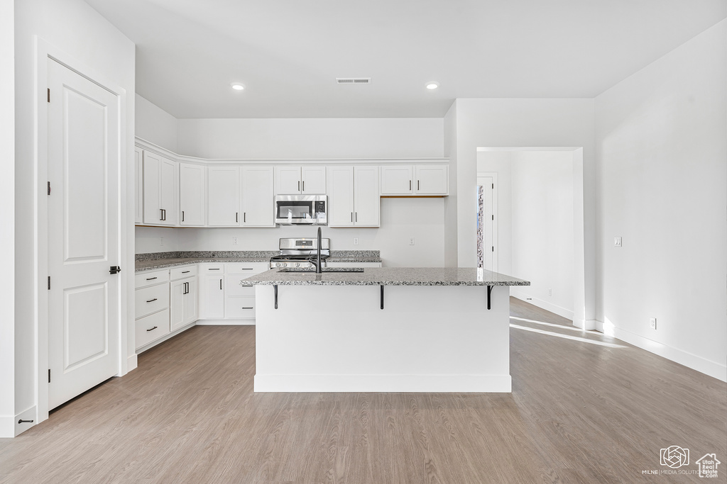 Kitchen with stainless steel appliances, light stone counters, a kitchen island with sink, light wood finished floors, and white cabinetry