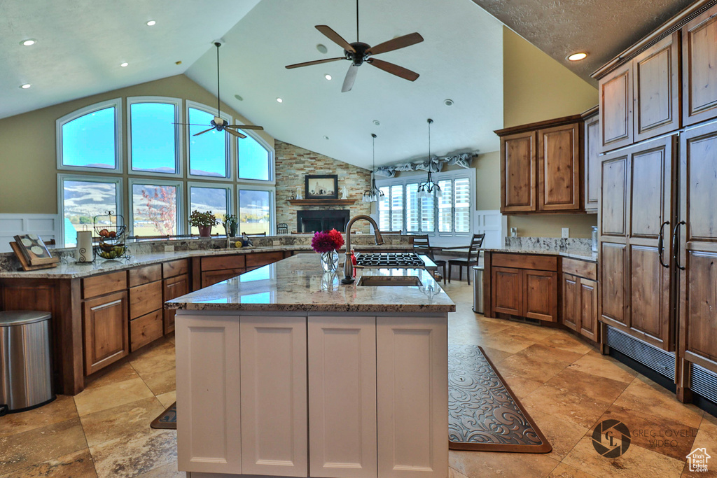 Kitchen with a ceiling fan, a stone fireplace, a kitchen island with sink, high vaulted ceiling, and light stone countertops
