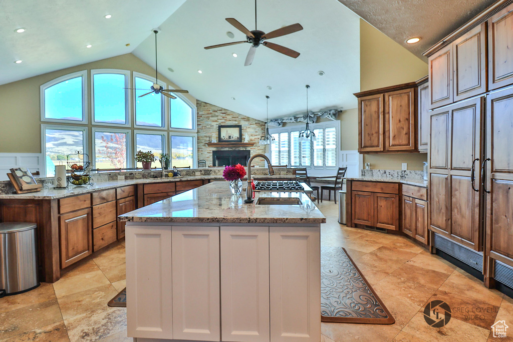 Kitchen with a ceiling fan, a kitchen island with sink, a stone fireplace, high vaulted ceiling, and brown cabinets