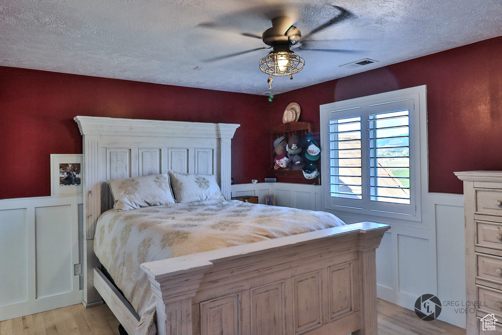Bedroom with light wood-style floors, a wainscoted wall, a textured ceiling, ceiling fan, and a decorative wall