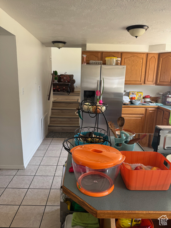 Kitchen featuring stainless steel refrigerator with ice dispenser, light tile patterned floors, brown cabinetry, a textured ceiling, and baseboards