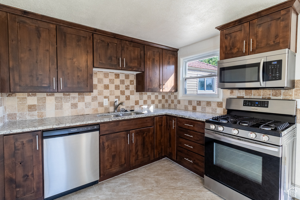 Kitchen featuring stainless steel appliances, a sink, light stone countertops, dark brown cabinets, and tasteful backsplash