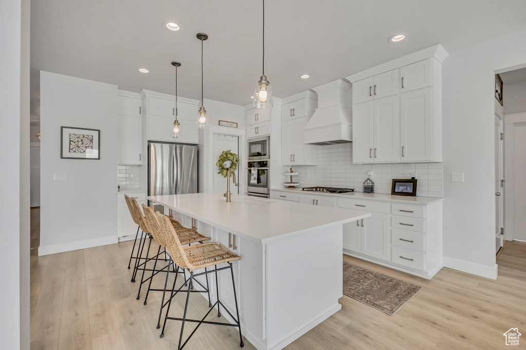Kitchen featuring custom range hood, appliances with stainless steel finishes, a breakfast bar, backsplash, and light wood-style flooring