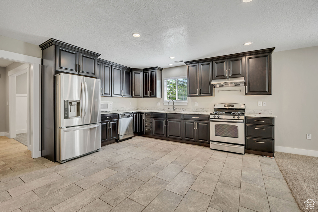 Kitchen with stainless steel appliances, under cabinet range hood, a textured ceiling, a sink, and baseboards