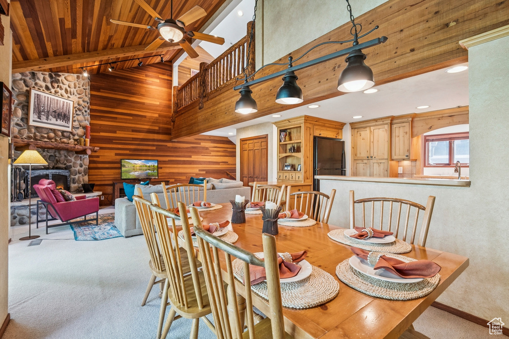 Carpeted dining room featuring a towering ceiling, ceiling fan, wood walls, wood ceiling with exposed beams, and a stone fireplace
