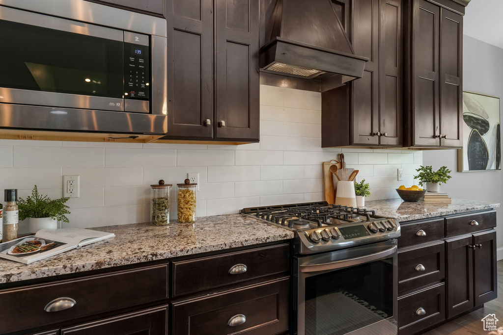 Kitchen with appliances with stainless steel finishes, premium range hood, dark brown cabinetry, light stone countertops, and decorative backsplash