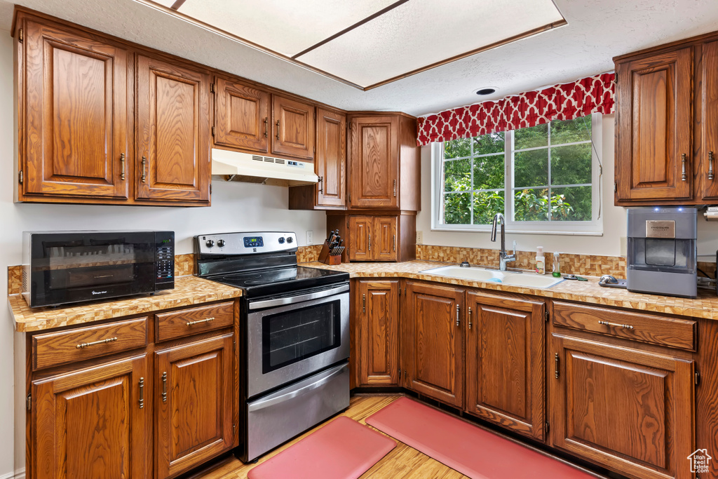 Kitchen featuring stainless steel range with electric stovetop, under cabinet range hood, a sink, brown cabinets, and light countertops