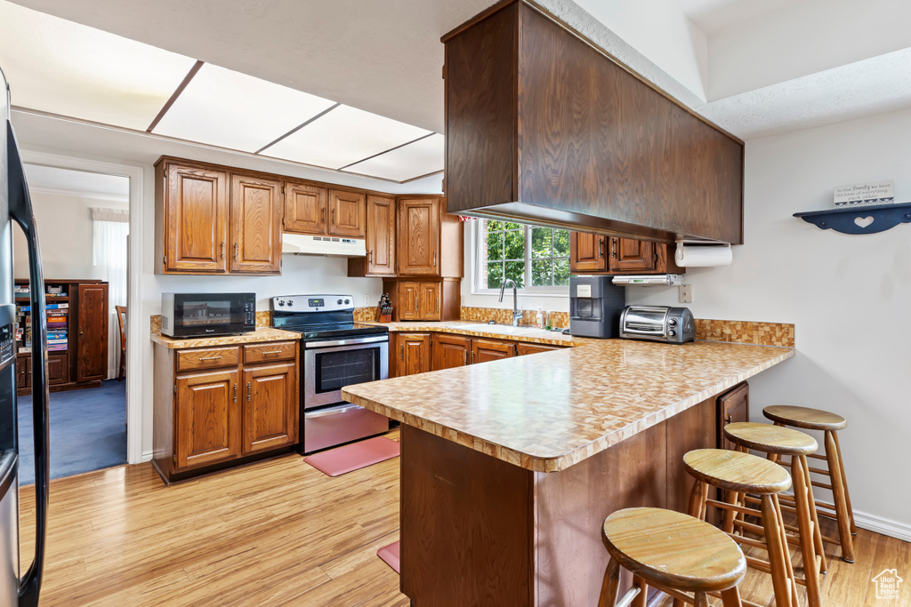 Kitchen with electric range, under cabinet range hood, a peninsula, light wood-type flooring, and a kitchen breakfast bar