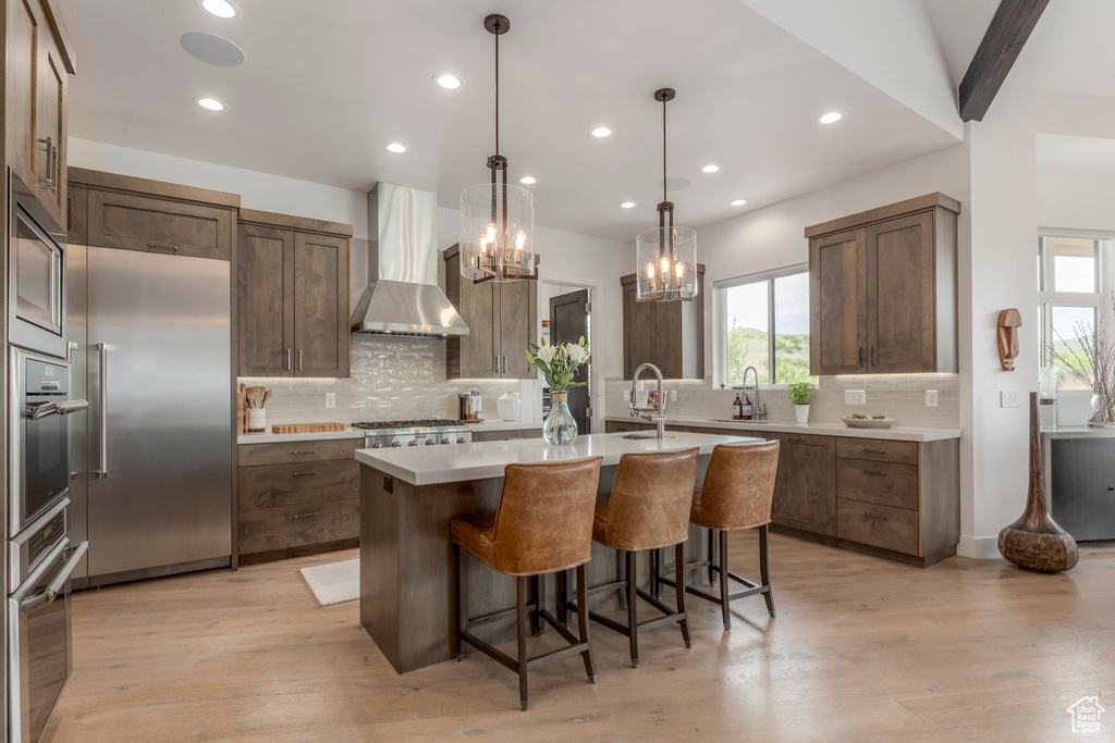 Kitchen featuring built in appliances, wall chimney range hood, a center island with sink, healthy amount of natural light, and recessed lighting
