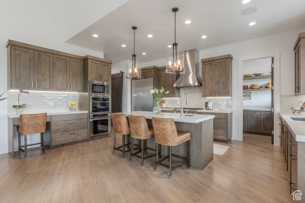 Kitchen featuring wall chimney exhaust hood, stainless steel appliances, decorative backsplash, an island with sink, and a breakfast bar area