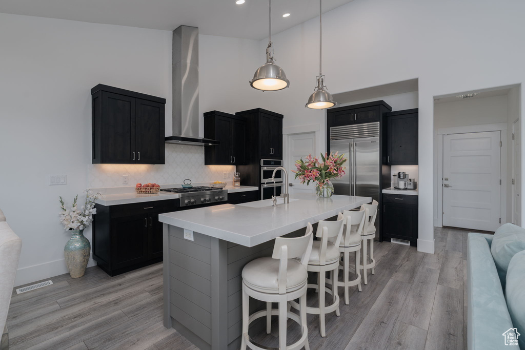 Kitchen featuring wall chimney exhaust hood, dark cabinets, a towering ceiling, stainless steel appliances, and light countertops