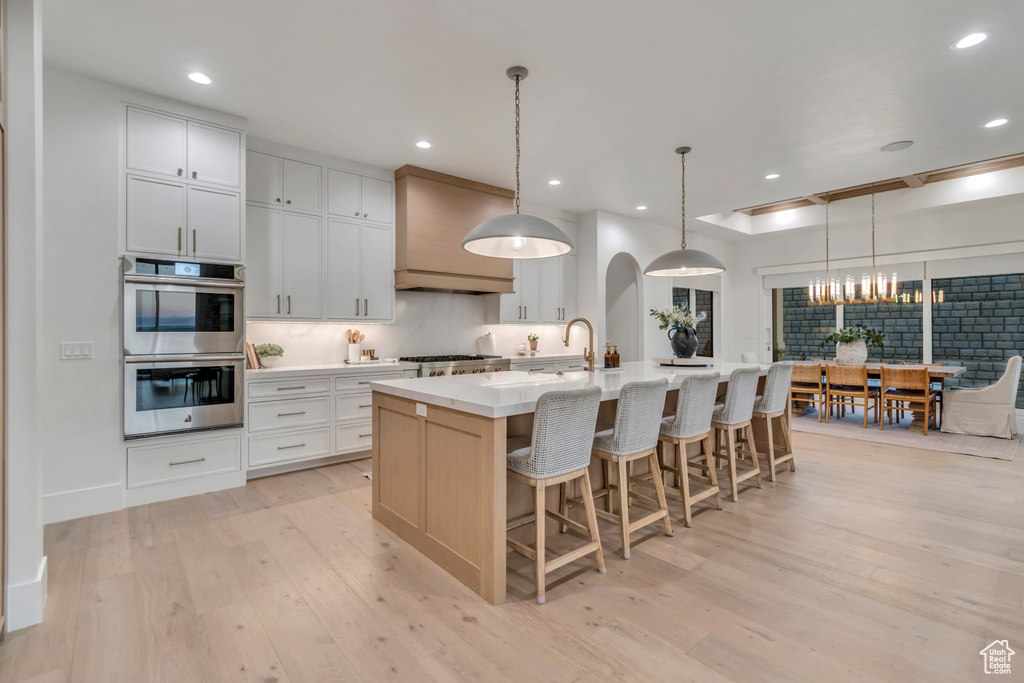 Kitchen featuring stainless steel double oven, light wood finished floors, a center island with sink, a breakfast bar area, and recessed lighting