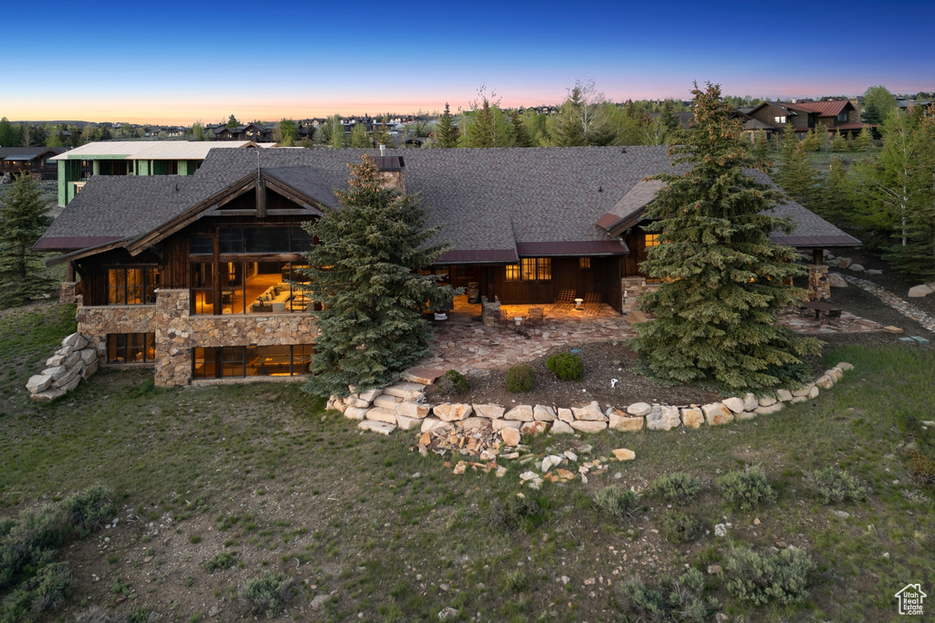 Back of house at dusk featuring stone siding, roof with shingles, a chimney, and a lawn