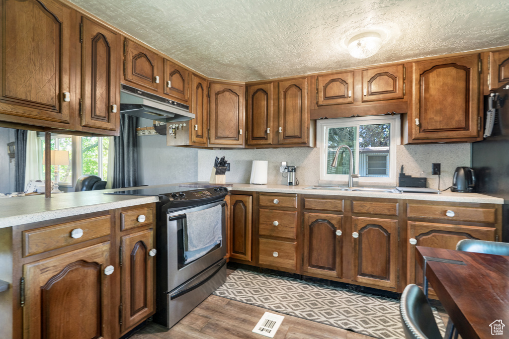 Kitchen with electric stove, under cabinet range hood, a sink, a textured ceiling, and brown cabinetry