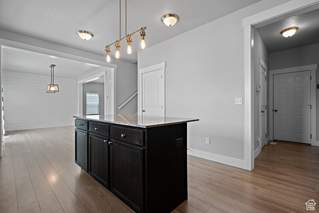 Kitchen featuring dark cabinetry, wood finished floors, light stone counters, decorative light fixtures, and a center island
