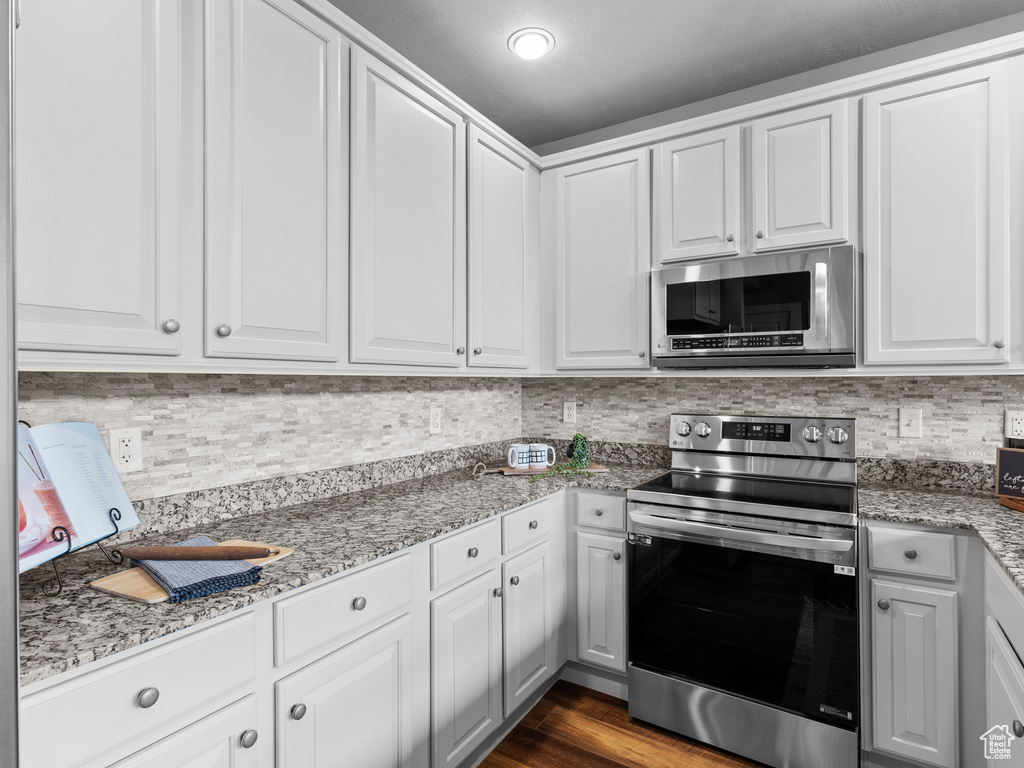 Kitchen with stainless steel appliances, decorative backsplash, white cabinets, light stone counters, and dark wood finished floors