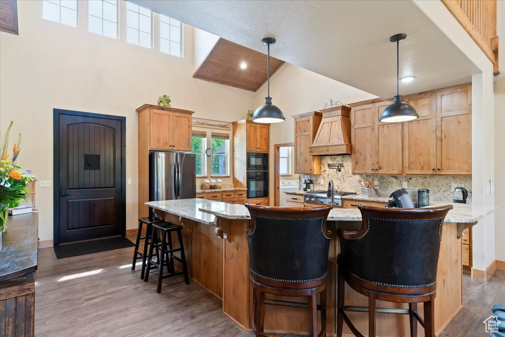 Kitchen featuring custom range hood, high vaulted ceiling, black appliances, decorative backsplash, and wood finished floors