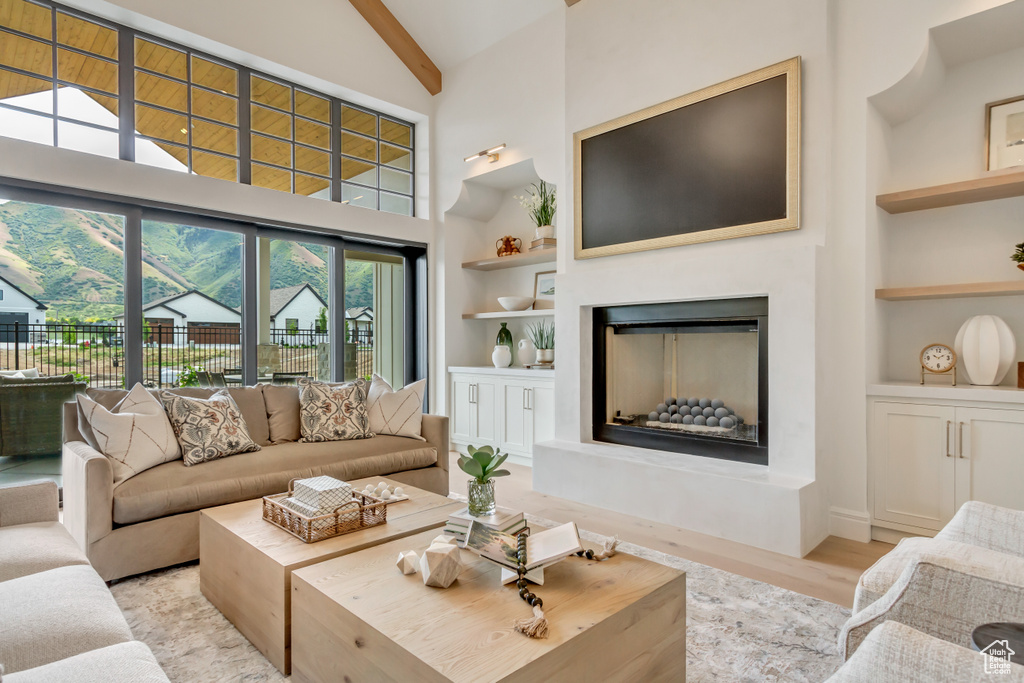 Living room featuring built in features, high vaulted ceiling, a fireplace with raised hearth, beam ceiling, and light wood-style flooring