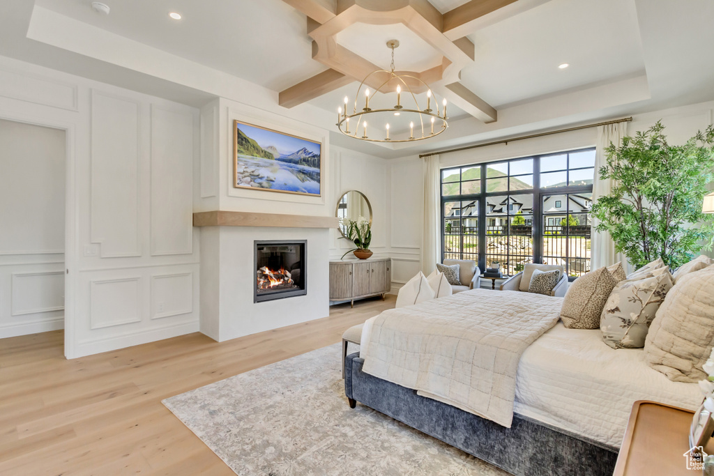 Bedroom featuring a decorative wall, a chandelier, light wood-style flooring, coffered ceiling, and a glass covered fireplace