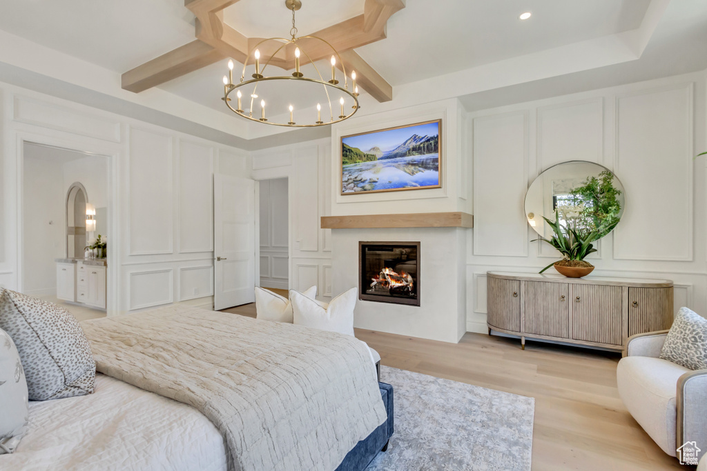 Bedroom with a decorative wall, light wood-type flooring, a chandelier, a glass covered fireplace, and beamed ceiling