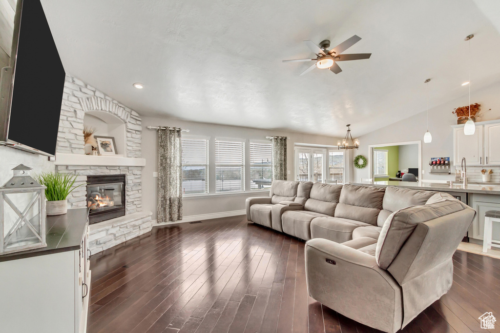 Living area featuring lofted ceiling, ceiling fan, a stone fireplace, dark wood finished floors, and a chandelier