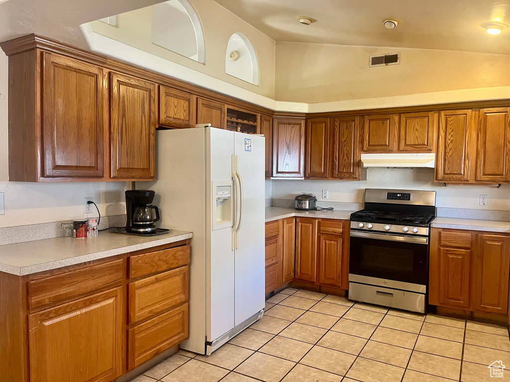 Kitchen featuring gas range, under cabinet range hood, light tile patterned flooring, white fridge with ice dispenser, and brown cabinets