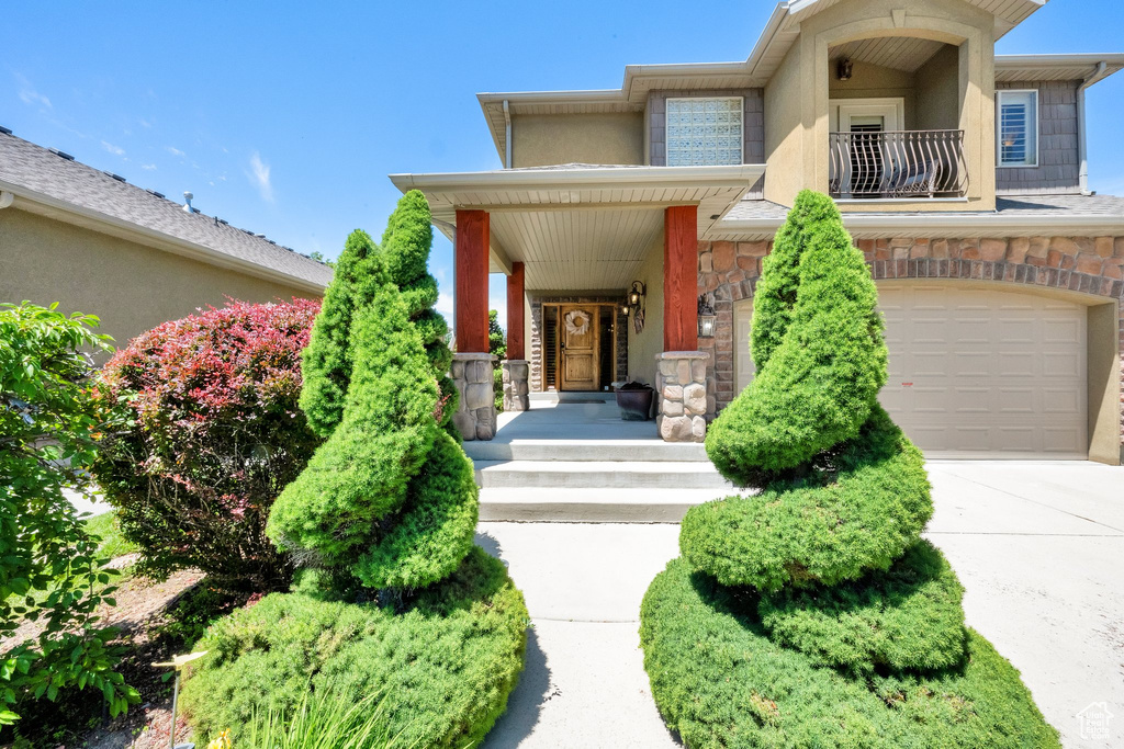 View of exterior entry with an attached garage, stone siding, a balcony, driveway, and stucco siding