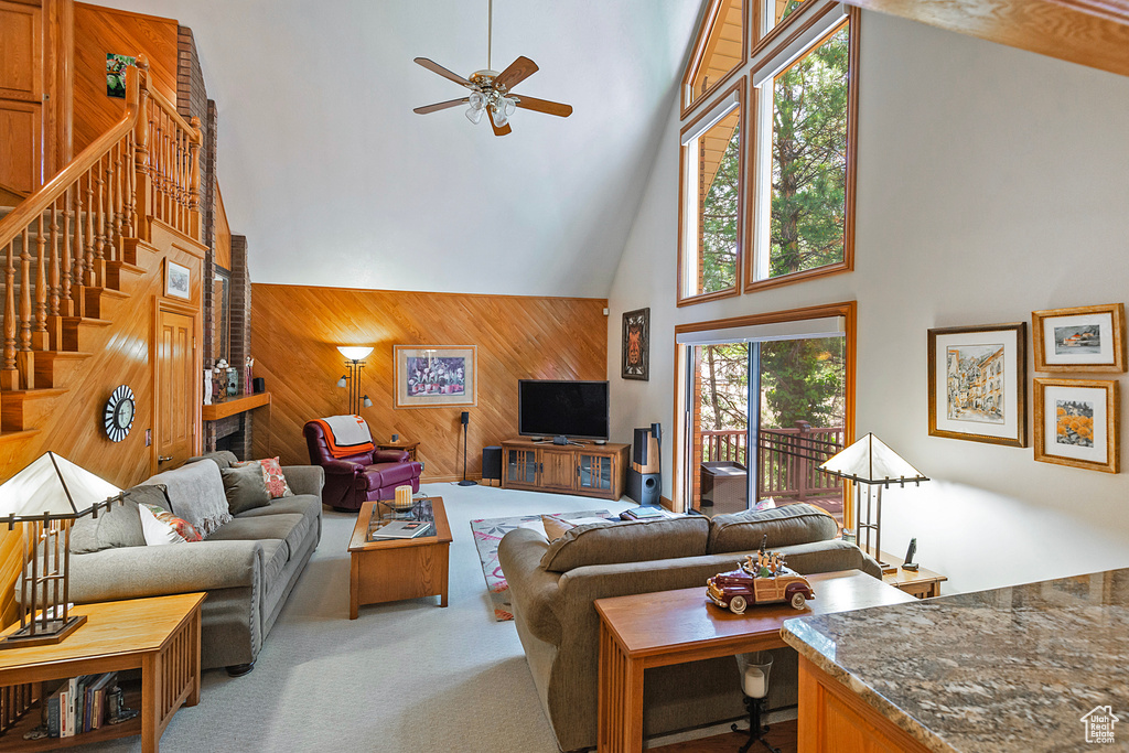 Living area featuring a high ceiling, a ceiling fan, wooden walls, stairway, and light colored carpet