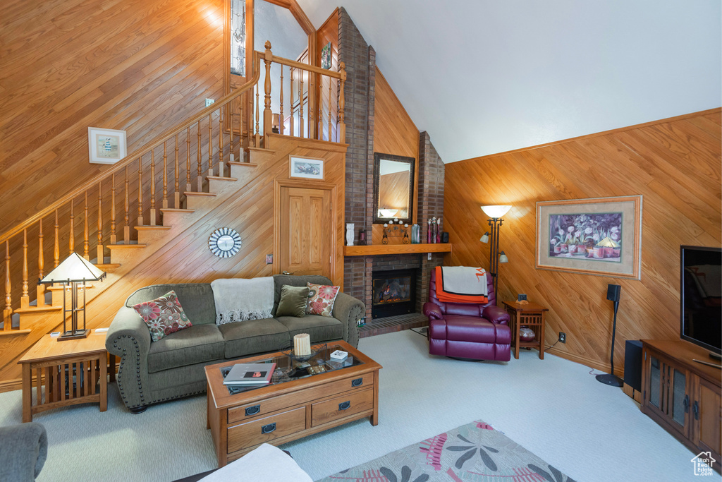 Carpeted living room with wooden walls, high vaulted ceiling, stairs, and a brick fireplace