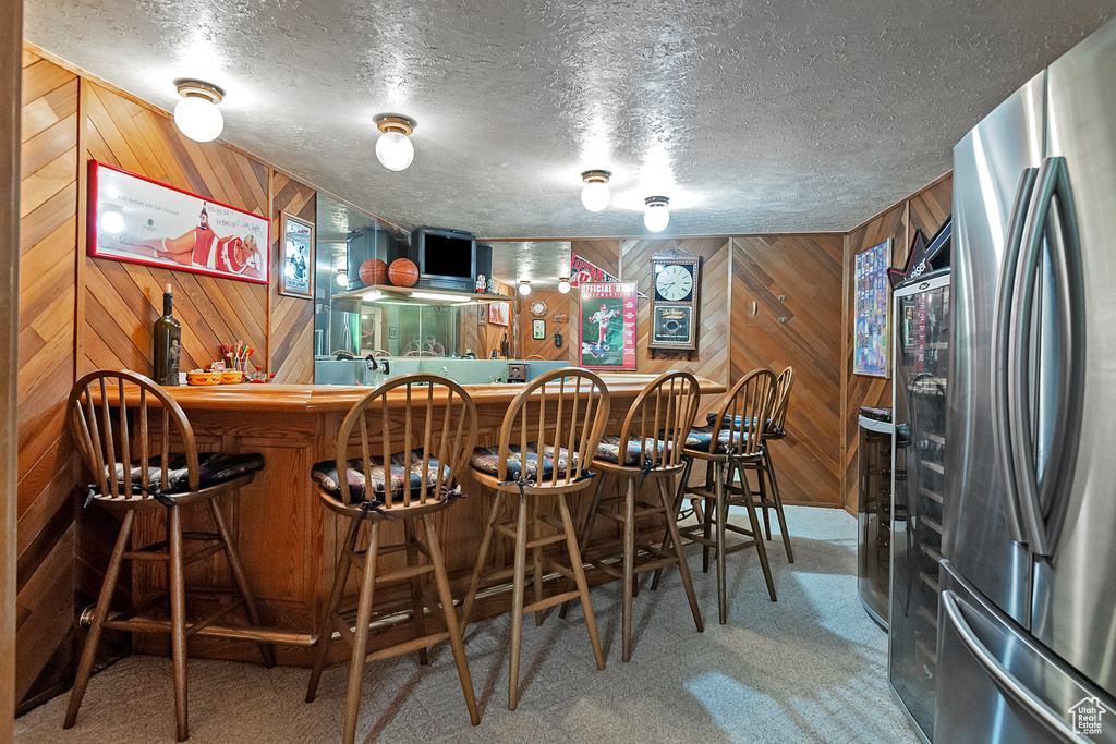 Indoor bar featuring freestanding refrigerator, carpet floors, wood walls, and a textured ceiling