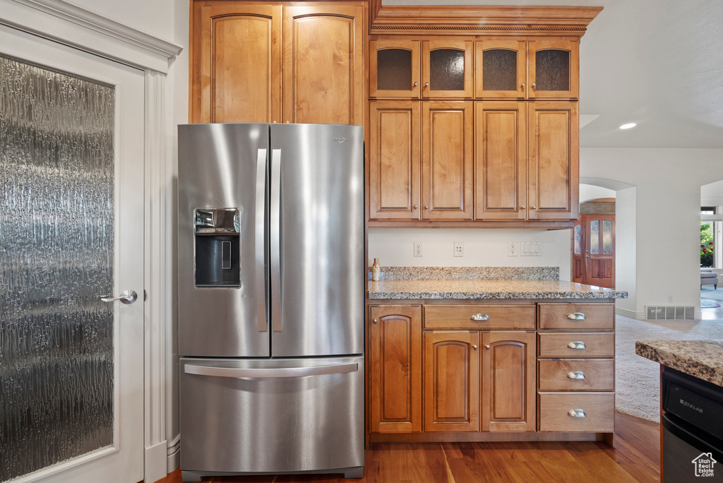 Kitchen featuring stainless steel fridge, arched walkways, brown cabinets, glass insert cabinets, and light stone counters
