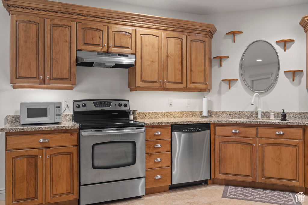 Kitchen with stainless steel appliances, under cabinet range hood, brown cabinets, light stone counters, and a sink