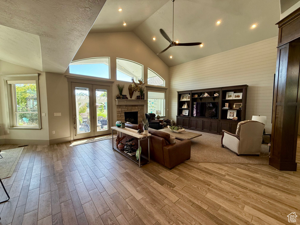 Living room with a stone fireplace, light wood-style flooring, ceiling fan, high vaulted ceiling, and baseboards