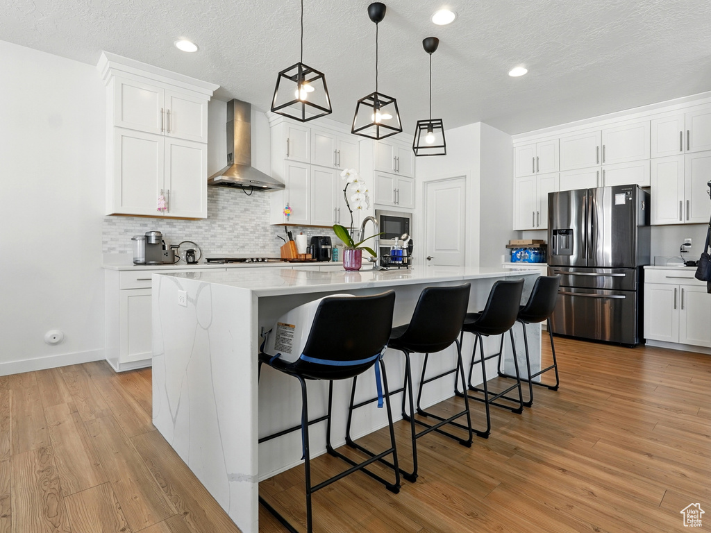 Kitchen with wall chimney exhaust hood, stainless steel fridge, light countertops, tasteful backsplash, and an island with sink