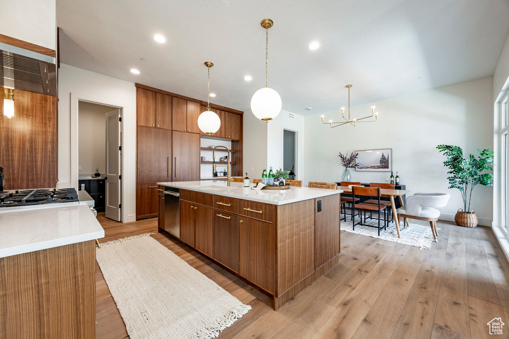 Kitchen with brown cabinetry, a chandelier, an island with sink, dishwasher, and light countertops